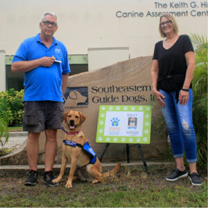 Dave and Cheryl Poage stand outside with Sully in front of a large rock that has 'Southeastern Guide Dogs' along with their logo engraved in it.