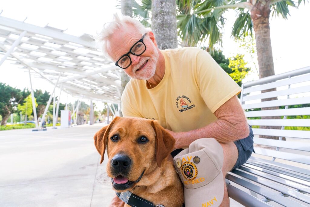 Ed and Sully, a yellow lab guide dog, sit outside in Florida on a metal bench.