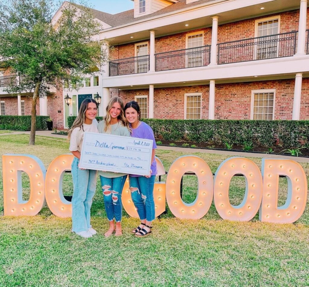 Three DG members holding check and standing in front of DO GOOD sign and house