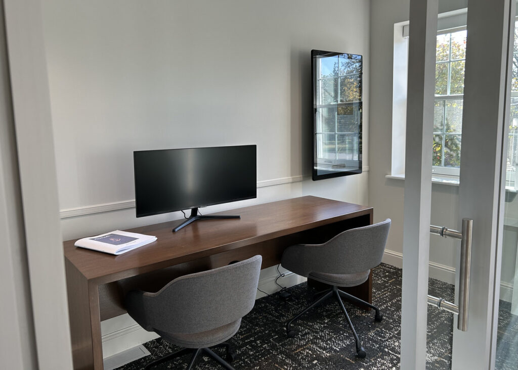 A small room with a glass door that has a wooden desk, a monitor, and two chairs. A vertical screen is to the right of the desk on the wall.