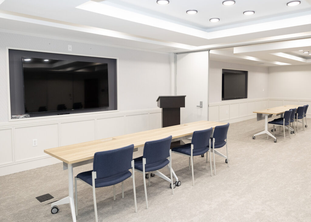 Two long tables with chairs sit in the front of a room. There is a podium and two screens mounted on the wall.