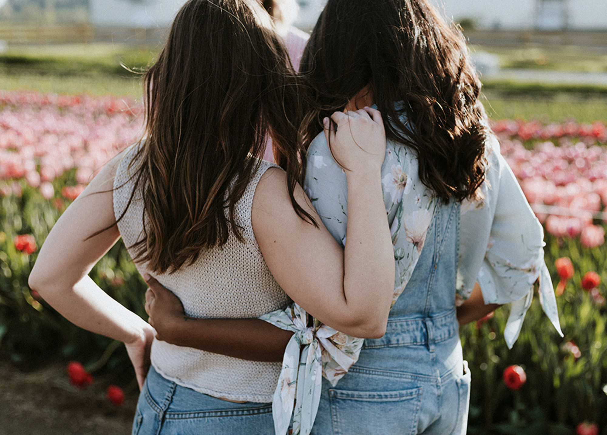 Two women stand in a field facing away from the camera with their arms around one another's back.