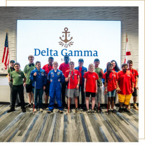 A group of 15 space camp attendees stand in front of a screen with the Delta Gamma logo on it.