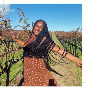 Rhyca is smiling at the camera with her arms spread out at her sides. She is standing outside surrounded by grape vines.