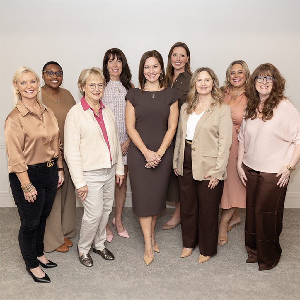 Nine women stand in a group indoors in front of a white wall.